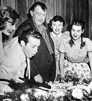 Andy Devine and his wife Dorothy (at Guy’s left) look on as Guy goes to work blowing out candles on his 1954 birthday cake. 