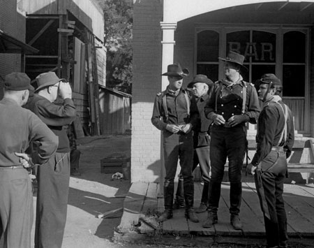 John Ford (L) sets up a scene for “The Horse Soldiers” in front of the Newton Station bar with William Holden, Willis Bouchey, John Wayne and unknown (William Wellman Jr.?)